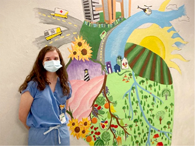 Young woman with long dark hair wearing blue scrubs and a face mask stands in front of a colorful mural on a tan wall