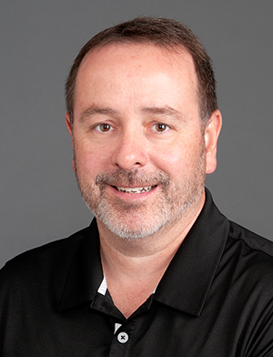 A man with dark hair and a goatee and wearing a black golf shirt smiles at the camera