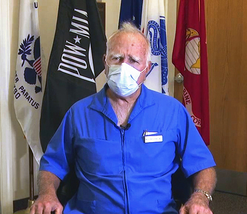 An older man in a bright blue shirt and face mask sits in front of various flags, including the Vietnam War Prisoner of War-Missing in Action one