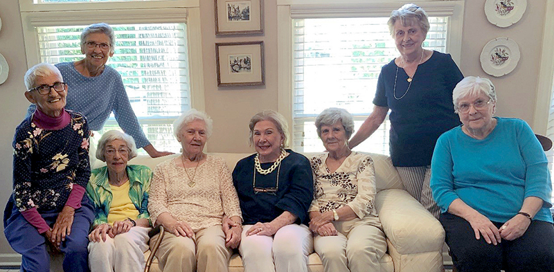 A group of eight older women sit together on a sofa in front of large windows