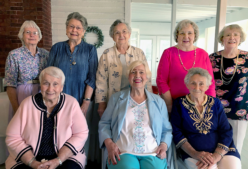 A group of eight older women sit and stand together in a sunroom