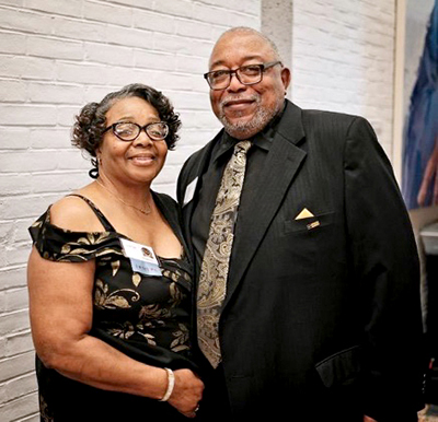 African American couple (man and woman) standing in front of white brick wall and wearing a suit and evening dress