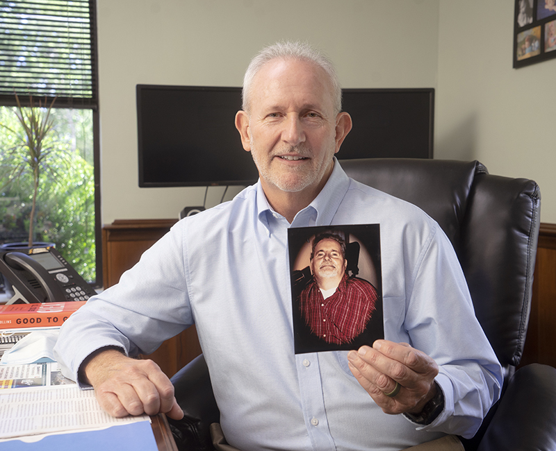 A white-haired man wearing a light blue button-down shirt sits at a desk and holds up a photo of another man