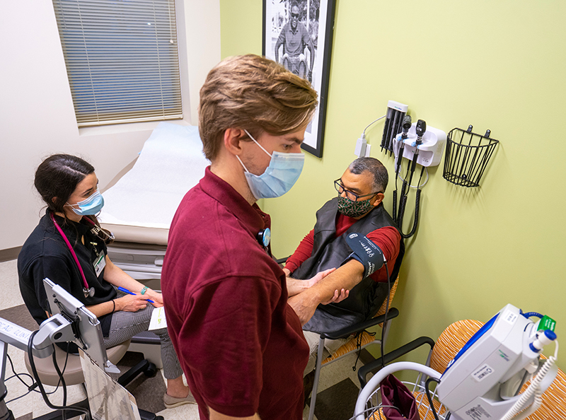 A student in a burgundy shirt and face mask takes the blood pressure of a male patient as a female student asks the patient questions