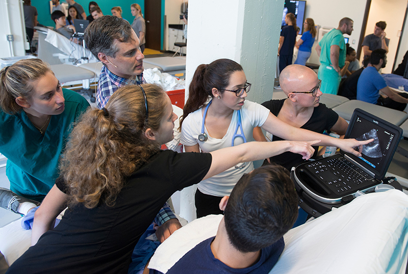 Two women point to an ultrasound monitor as male instructor, male patient and other students look