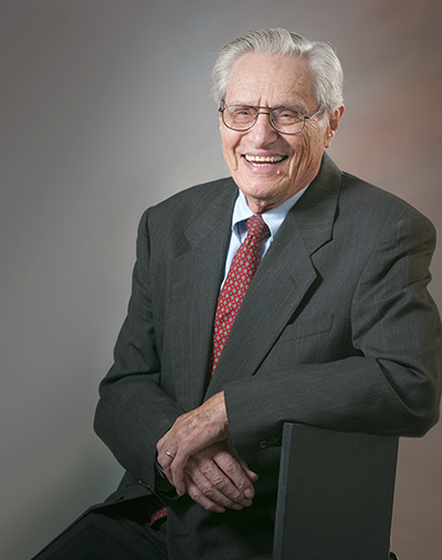 An older man with white hair and glasses smiles as he looks at the camera. he is wearing a suit and tie.