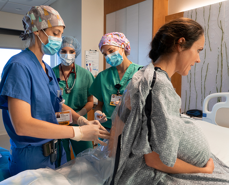 Two people in green scrubs watch as a person in blue scrubs places an epidural in the spine of an obstetric patient