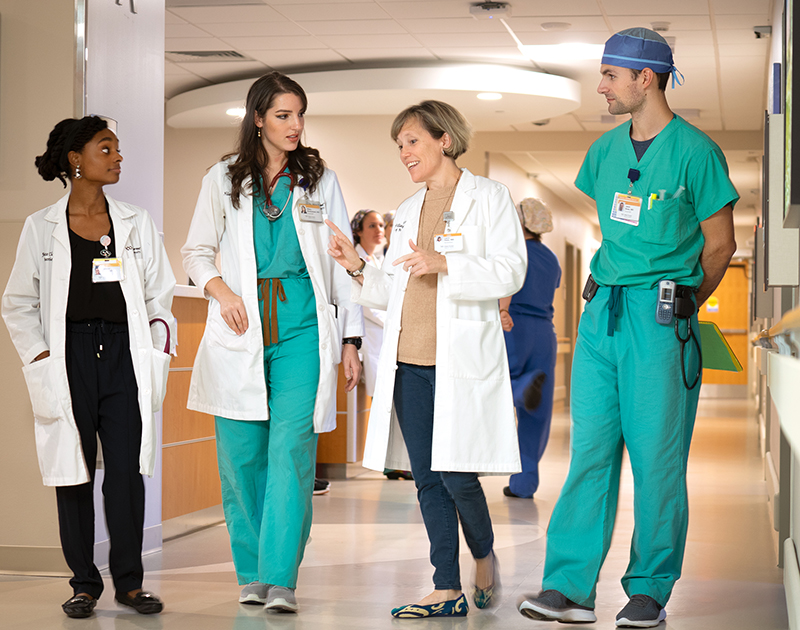 Four people in surgical scrubs and/or white coats walk down a hall while talking