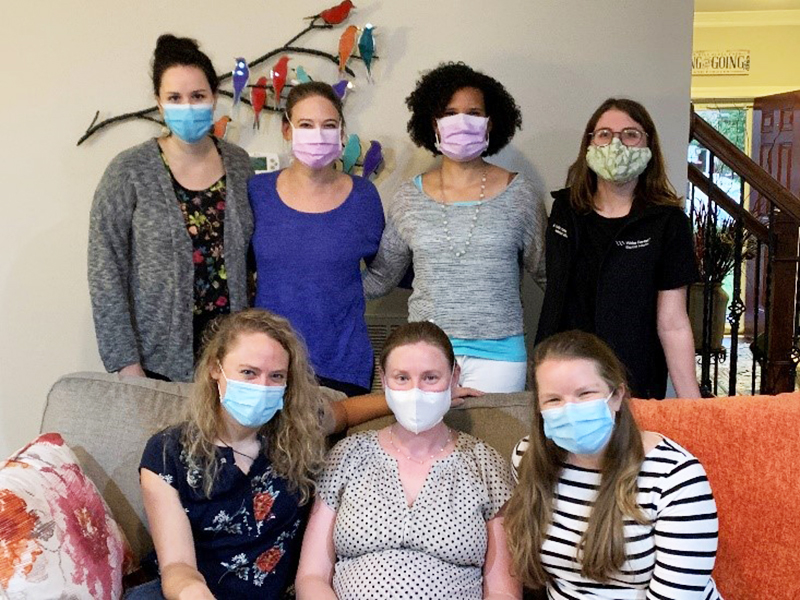 Seven women - three sitting on an orange couch and four standing behind - wear masks and pose for a group photo