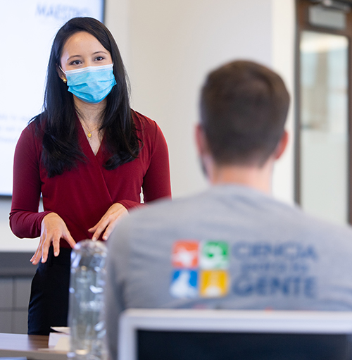 A woman of Asian descent wears a facial mask and burgundy shirt as she stands in front of a class and talks. 