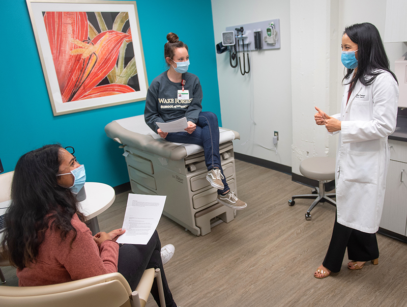 A woman of Asian descent wears a long white coat and facial mask as she talked to a young woman on an exam table and another one sitting in a chair in the exam room