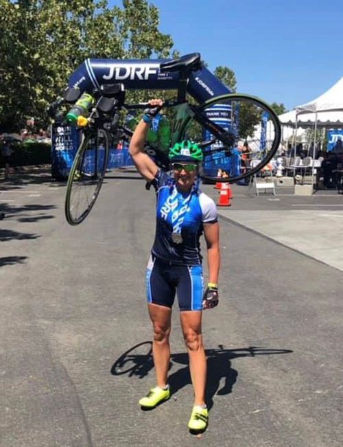 Tan, fit young woman in cycling clothes smiles and holds her bicycle up in one hand in front of an inflated arch with 'JDRF' on it