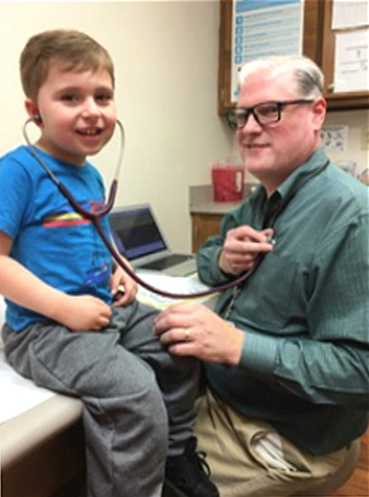 A blond man in a green shirt uses a stethoscope to listen to the heart of a young male patient sitting on an exam table wearing pants and a blue shirt