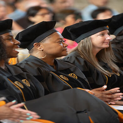 Graduating students sitting next to each other while wearing caps and gowns.